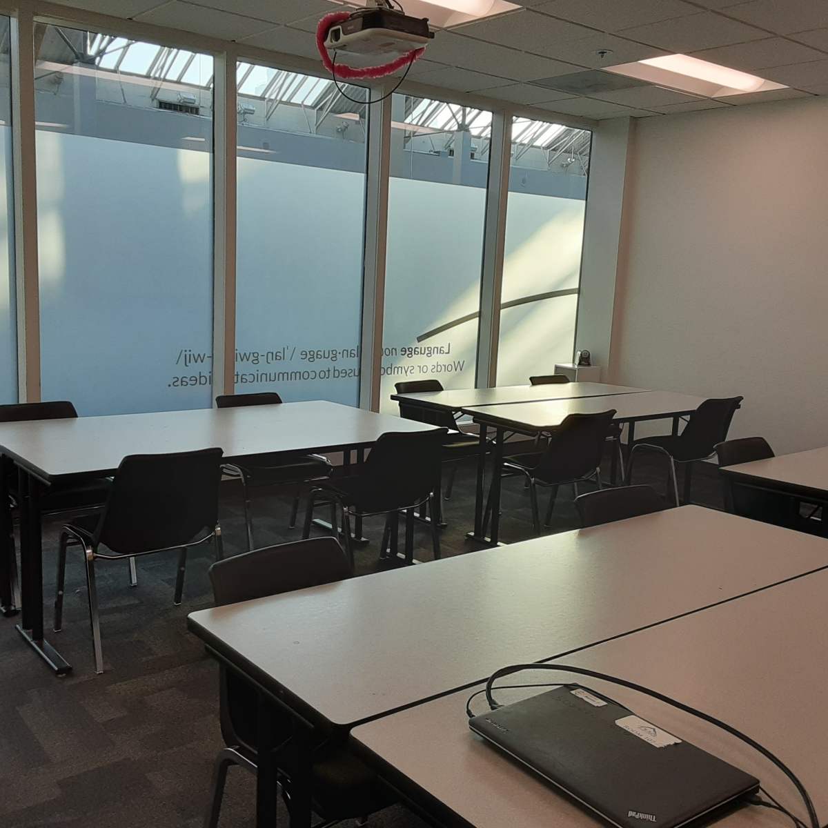 A well-lit classroom with multiple tables and chairs arranged in two groups, featuring large windows on one side.