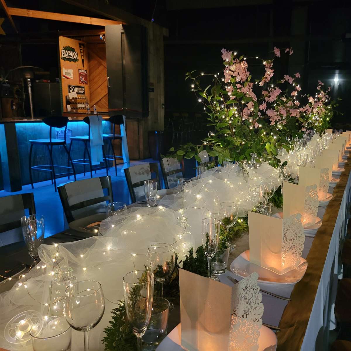 A long, elegantly set dining table with floral centerpieces and twinkling lights is arranged in a dimly lit space.