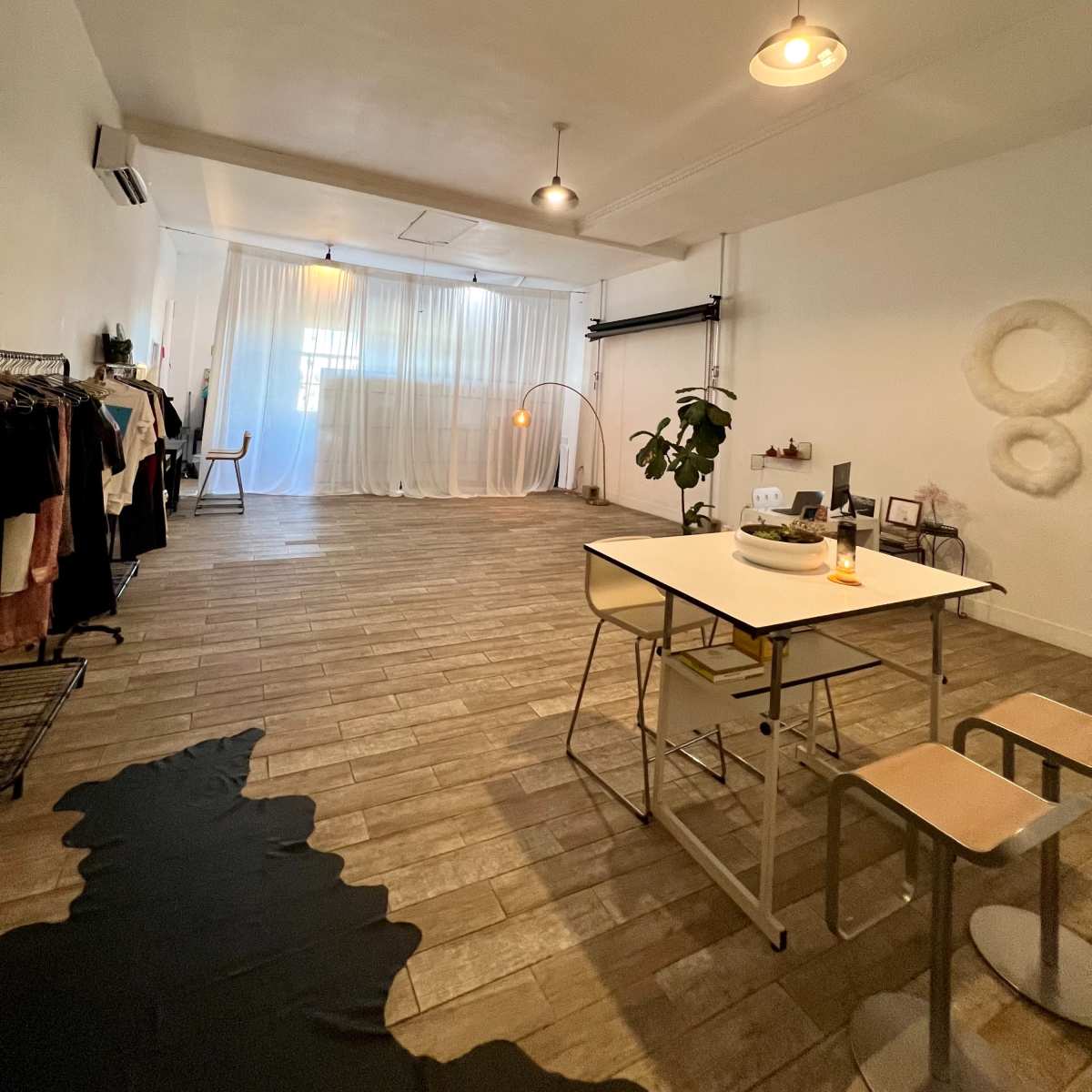 A spacious room with wooden flooring, a dining table with two chairs, and clothing racks along one wall, illuminated by soft lighting and curtains in the background.