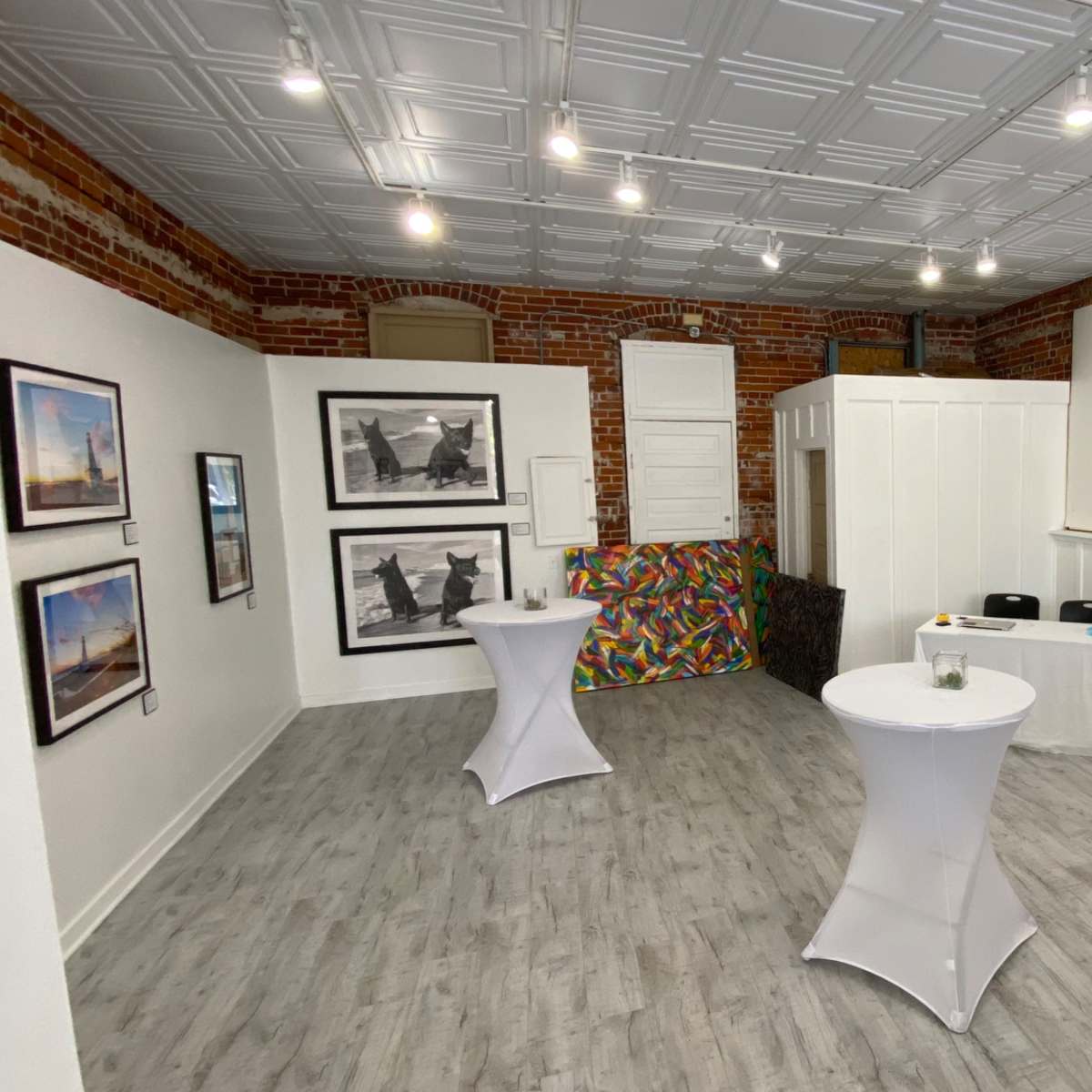 A spacious room with exposed brick walls, featuring artwork on display and two white cocktail tables set up for an event.