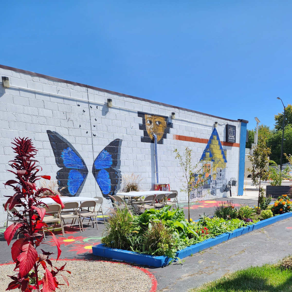 A colorful mural featuring a butterfly and a house decorates a wall in a garden setting with tables and greenery.