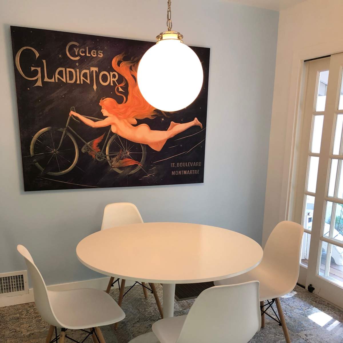 A round white dining table surrounded by four white chairs is positioned beneath a large vintage poster of a woman on a bicycle.