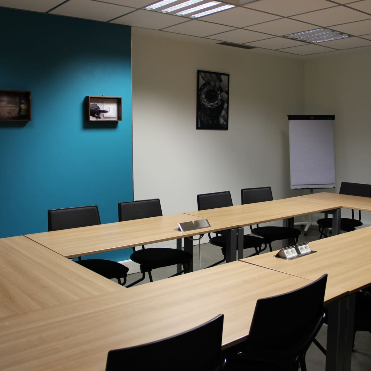 A conference room features a large rectangular table surrounded by black chairs, with teal and white walls, and wall-mounted framed images.