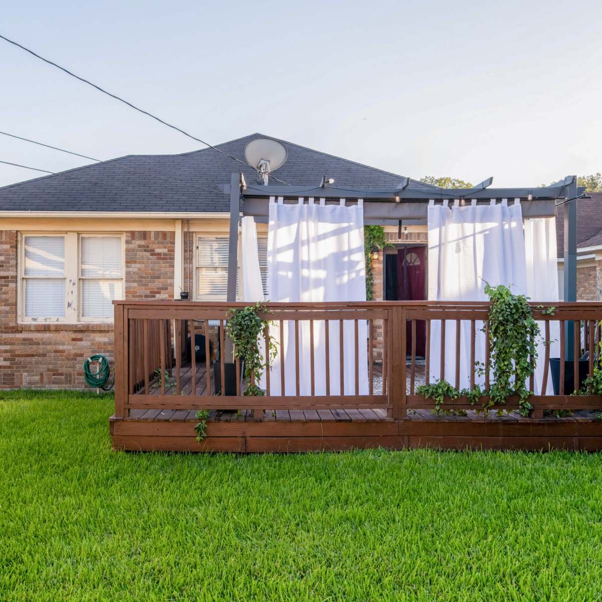 A brick house with a wooden deck is surrounded by green grass and features white curtains hanging from a pergola.