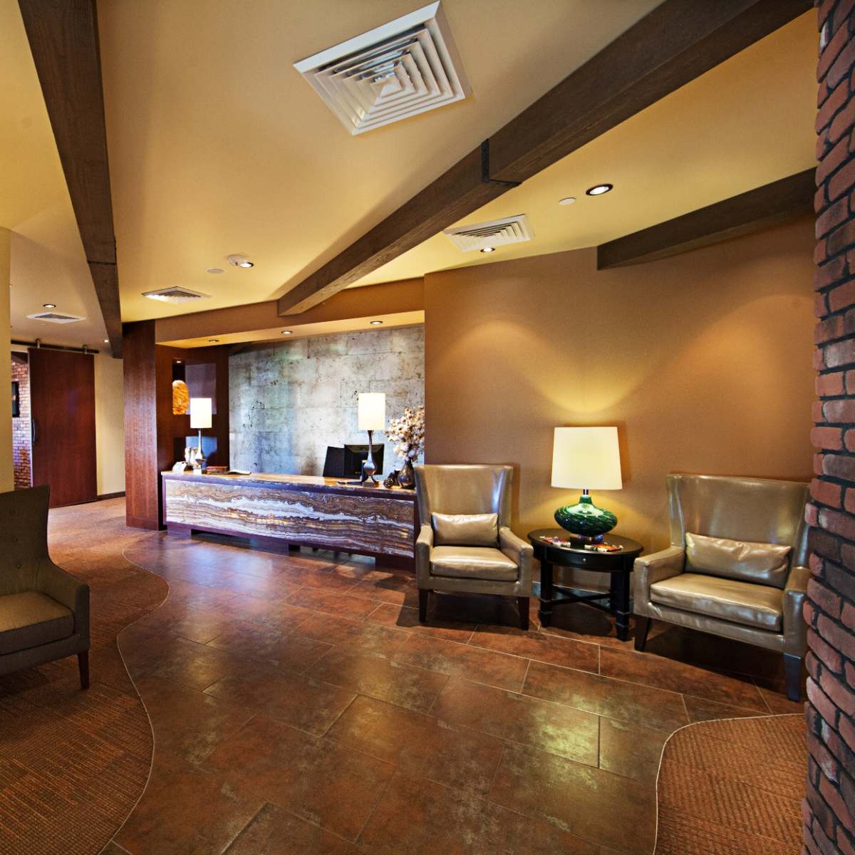 A lobby area with two armchairs, a side table, a reception desk, and decorative lamps against a warm-toned wall.