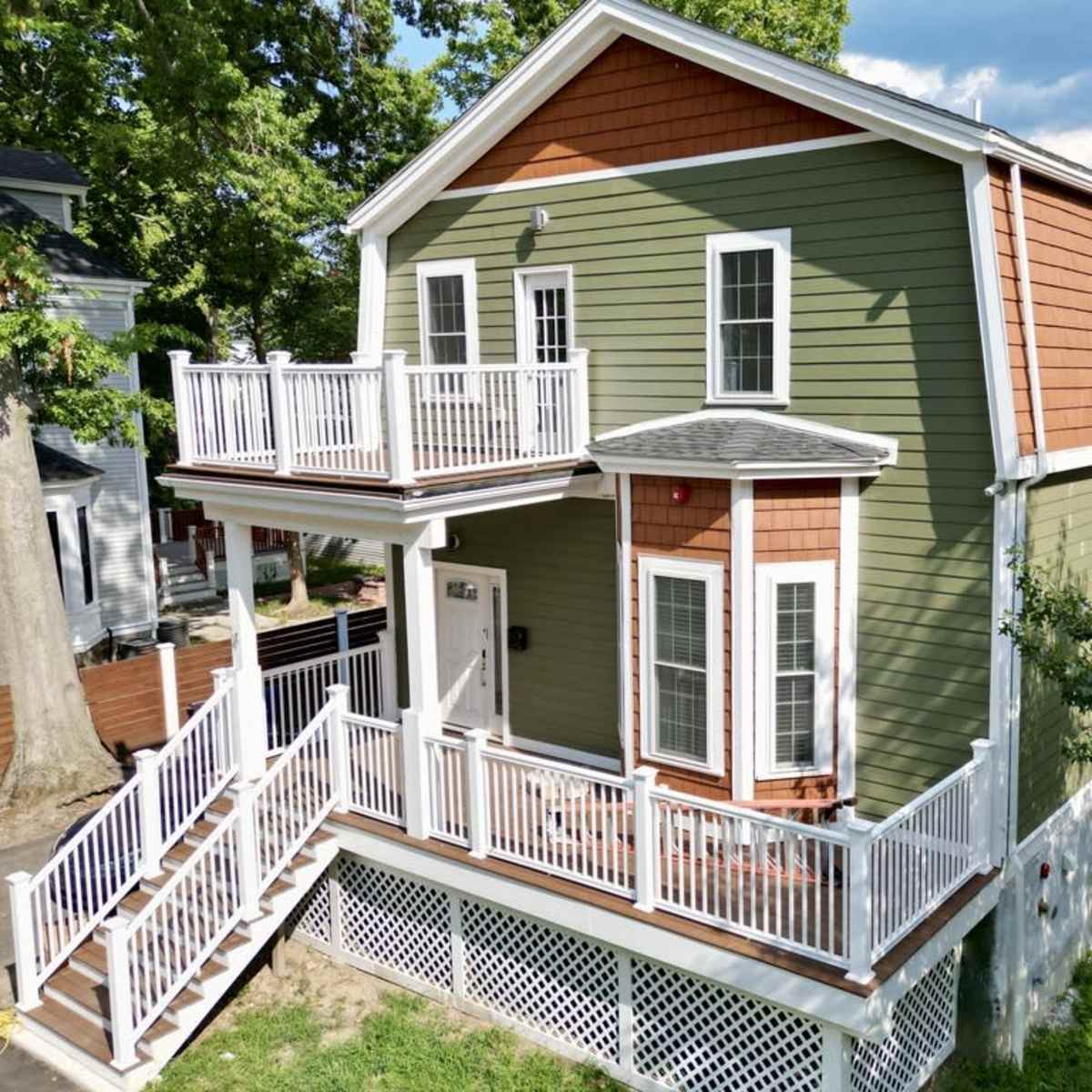 A two-story green house with a brown shingle roof, a front porch, and multiple white railings on the balconies.