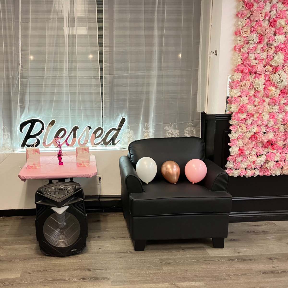 Two black chairs decorated with balloons, a pink table with a floral backdrop, and the word "Blessed" illuminated on the wall.