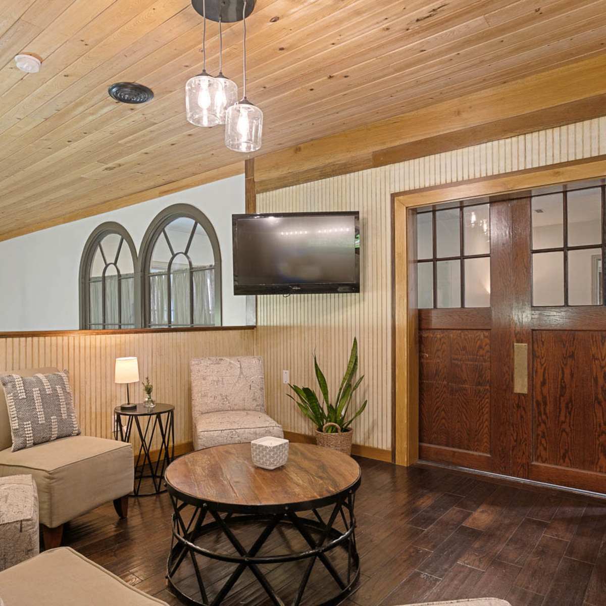 A living area with two armchairs, a circular coffee table, a flat-screen TV mounted on the wall, and a wooden front door with windows.