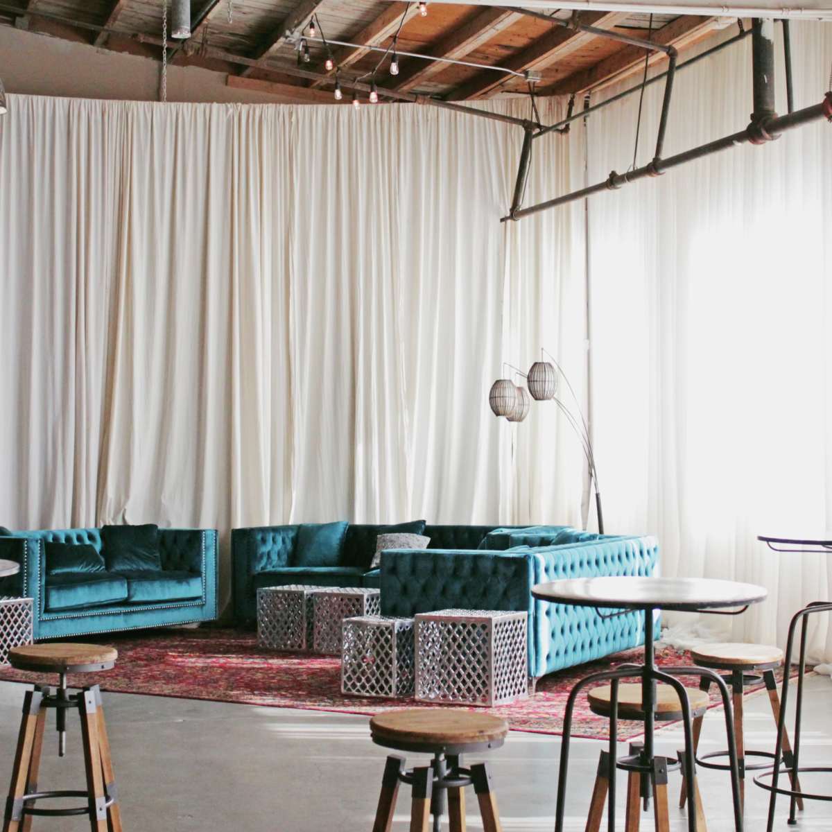 A spacious indoor area features turquoise couches, a patterned rug, and high tables with wooden stools against a backdrop of white drapes.
