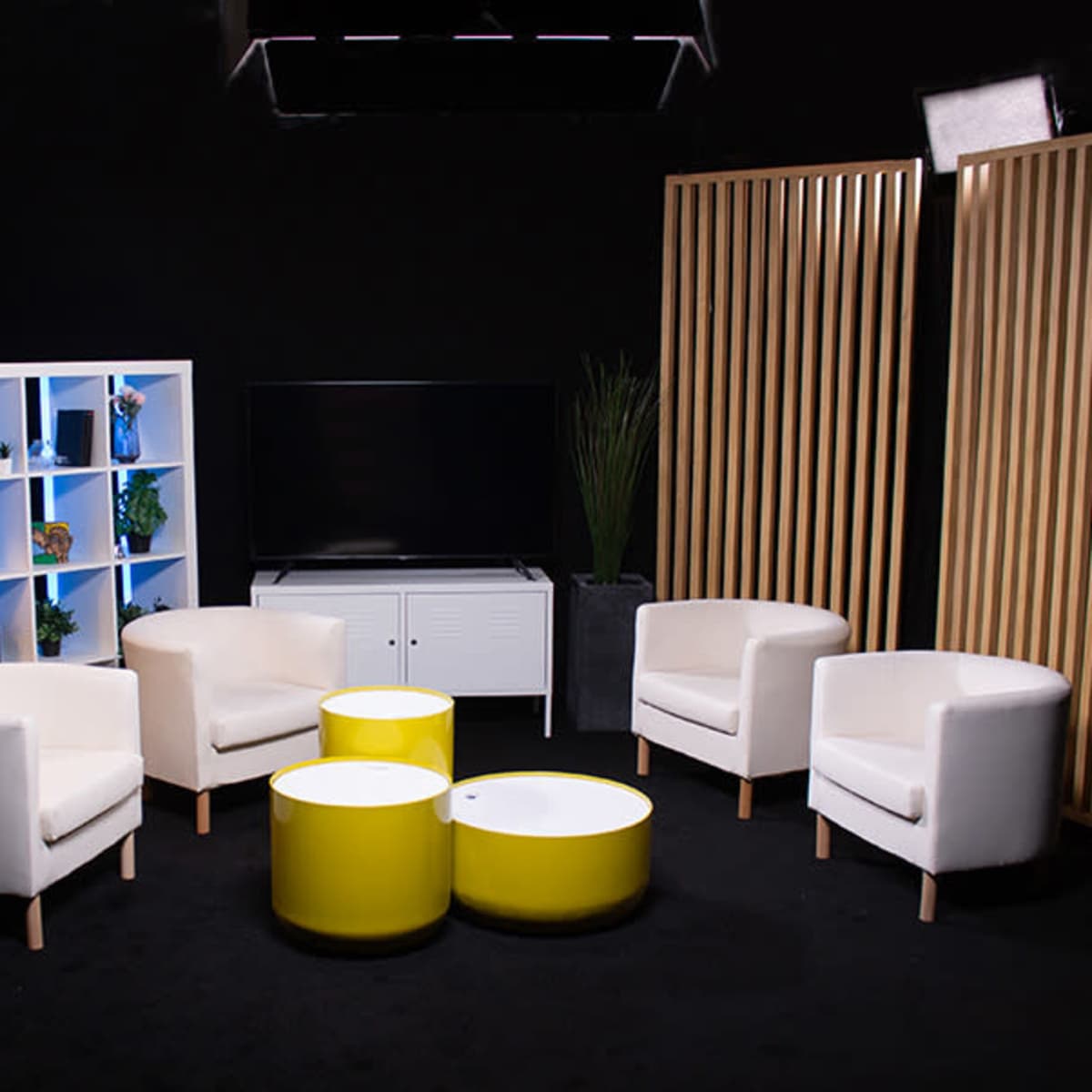 A modern studio setting with four white chairs arranged around two yellow coffee tables, a bookshelf, and a television on a low cabinet, all under soft lighting against a black backdrop.