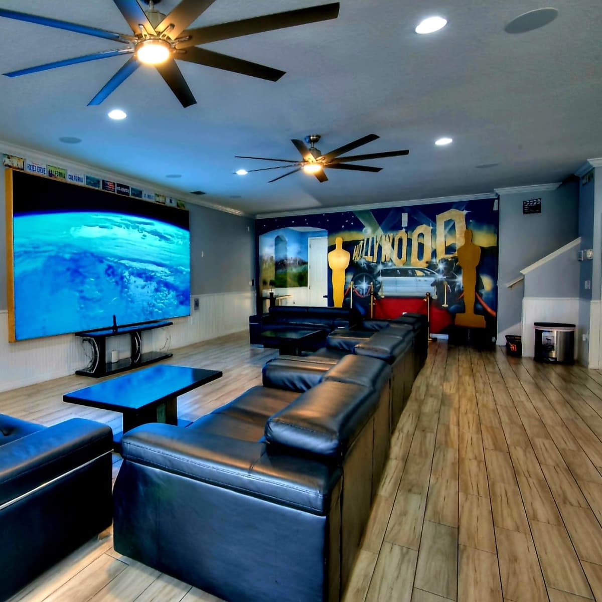 A large living room features black leather sofas, a central coffee table, and a wall-mounted screen displaying a scenic view, with decorative elements in a modern design.