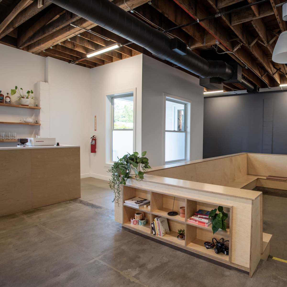 A modern office space with exposed wooden beams, minimalist furniture, and a low wooden seating area surrounded by large windows.