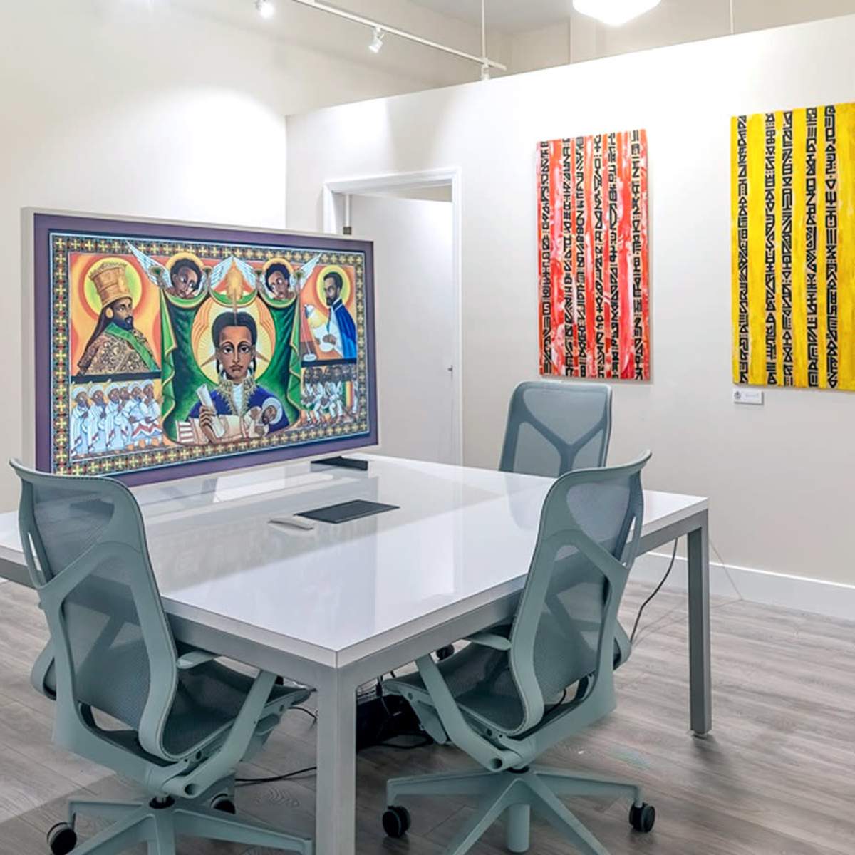 A modern gallery space featuring a central white table surrounded by chairs, with colorful artwork displayed on the walls behind it.