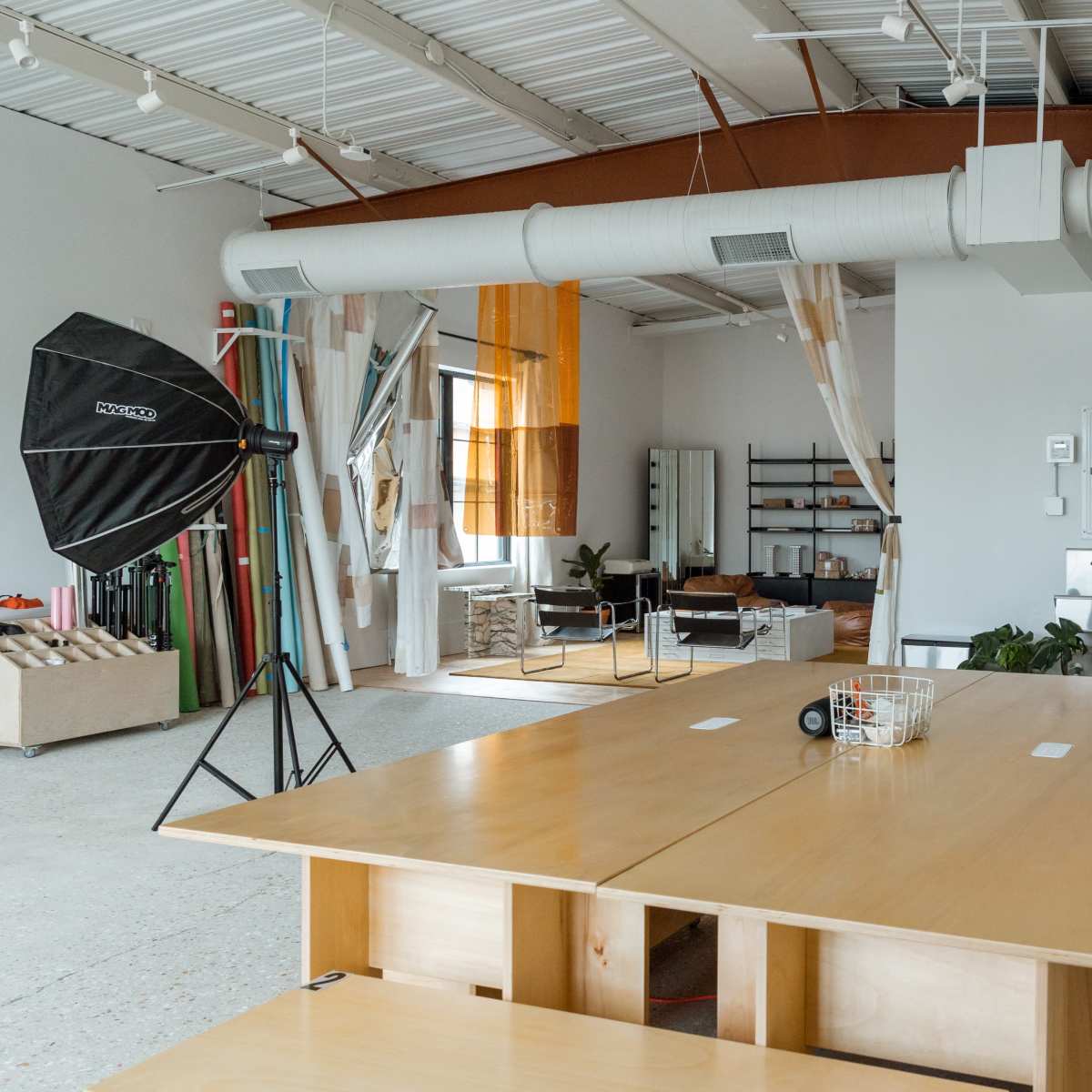 A spacious studio with a large wooden table, various lighting equipment, and a minimalist interior featuring shelves and couches.