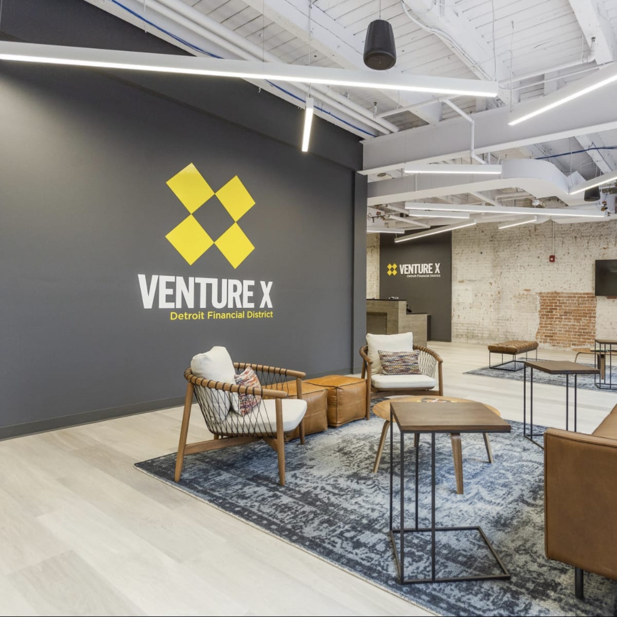 A modern office interior of Venture X, featuring a seating area with chairs and tables, a large wall logo, and exposed brick in the background.