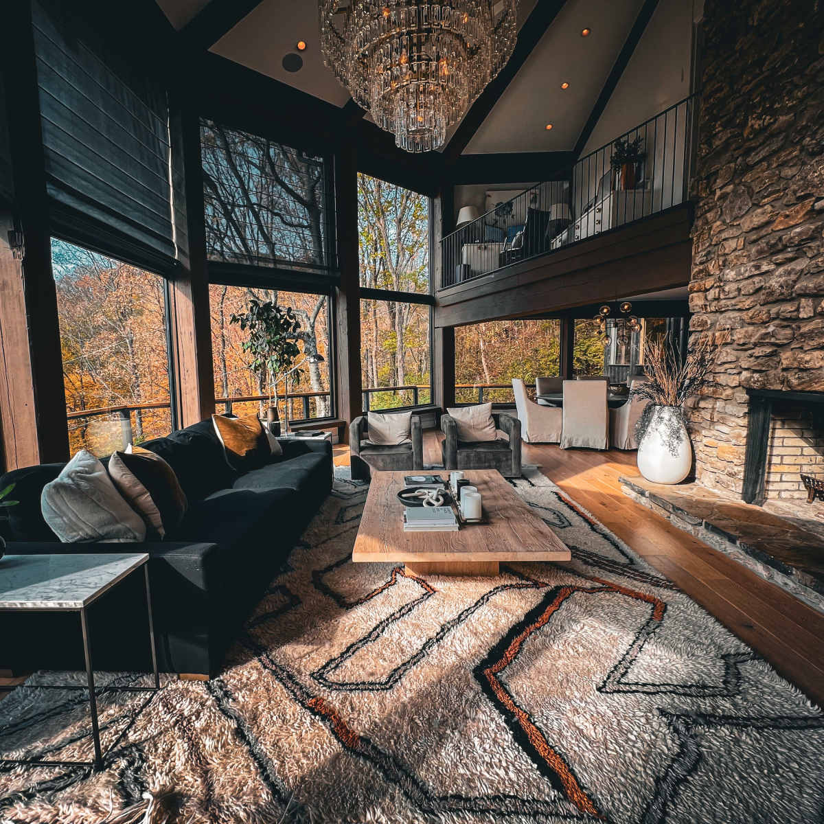A modern living room with large windows overlooking a forested area, featuring a dark sofa, a wooden coffee table, and a stone fireplace.