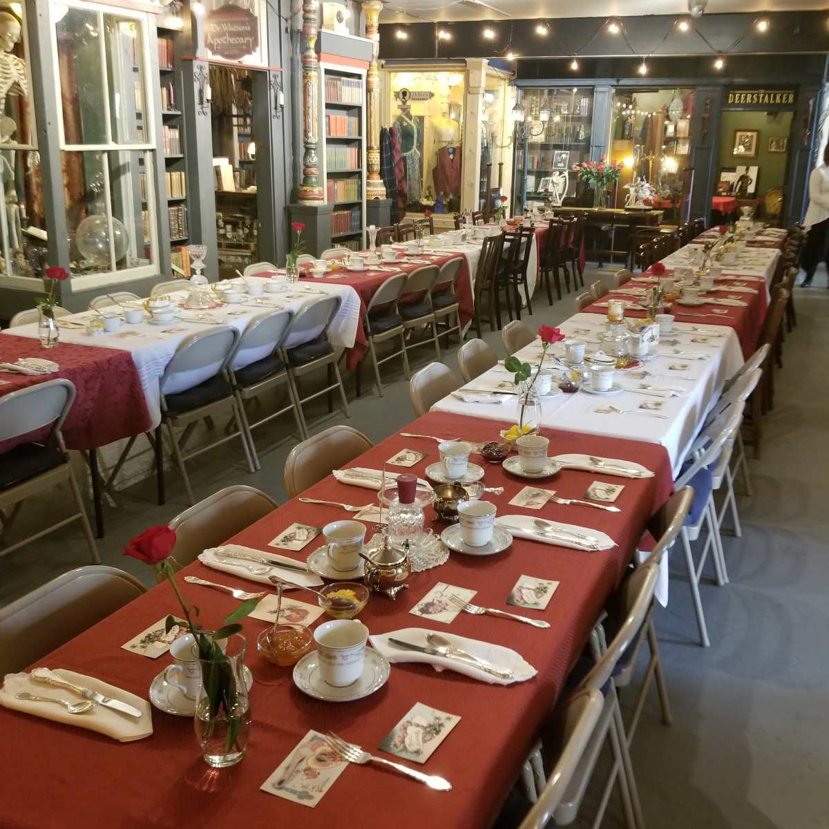 A long dining table is set with dishes, utensils, and flowers, surrounded by vintage decor and displays in a shop interior.