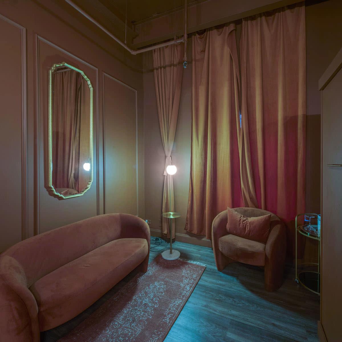 A small room featuring a curved sofa and armchair, a mirror, a floor lamp, and curtains in a warm color scheme.