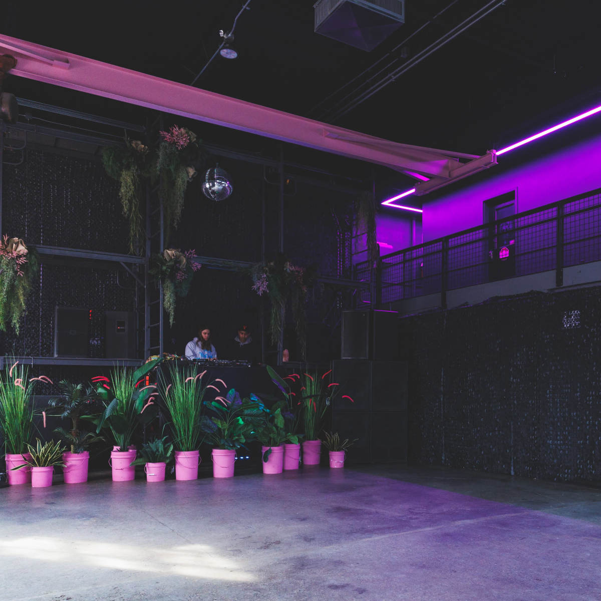 A modern indoor space with a dark interior, highlighted by purple neon lights, potted plants in pink containers, and a staircase leading to an upper level.