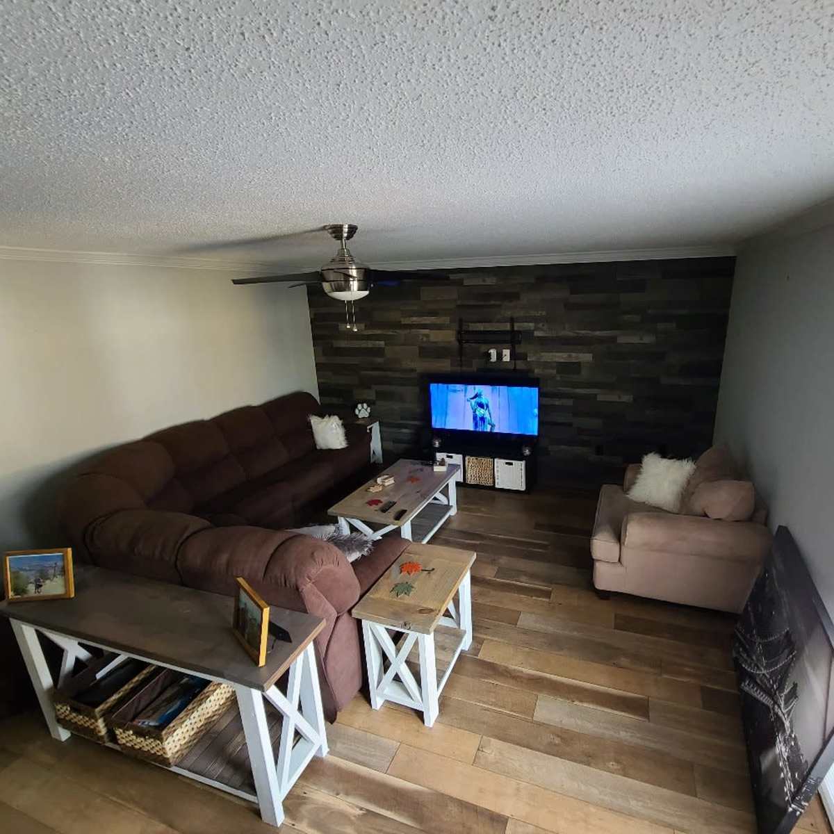 A living room features a brown sectional couch, two wooden tables, a television mounted on a stone accent wall, and a beige armchair.