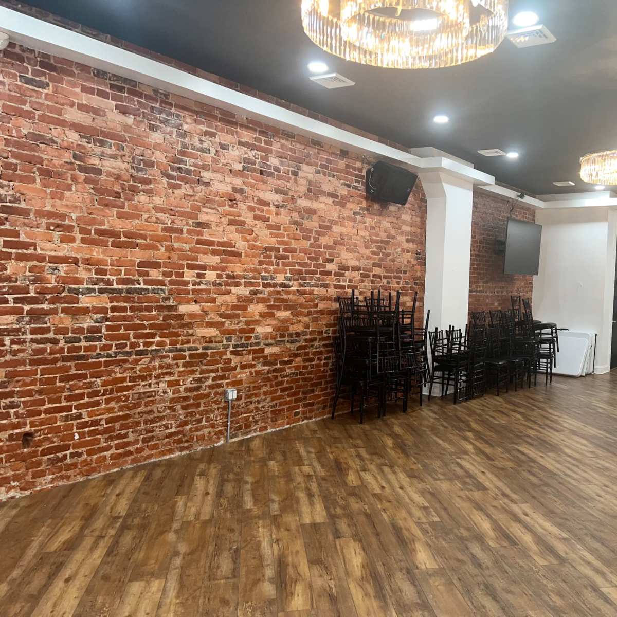 An empty room with exposed brick walls, wooden flooring, stacked black chairs, and decorative chandeliers.