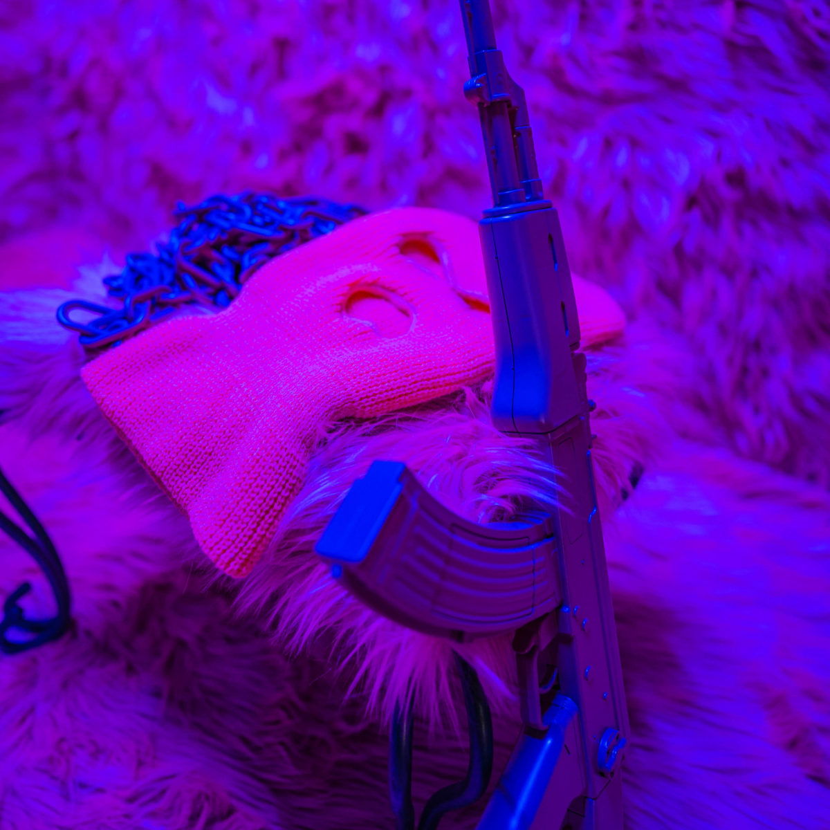 A model of an AK-47 rifle leans against a bright pink ski mask on a fuzzy purple background.