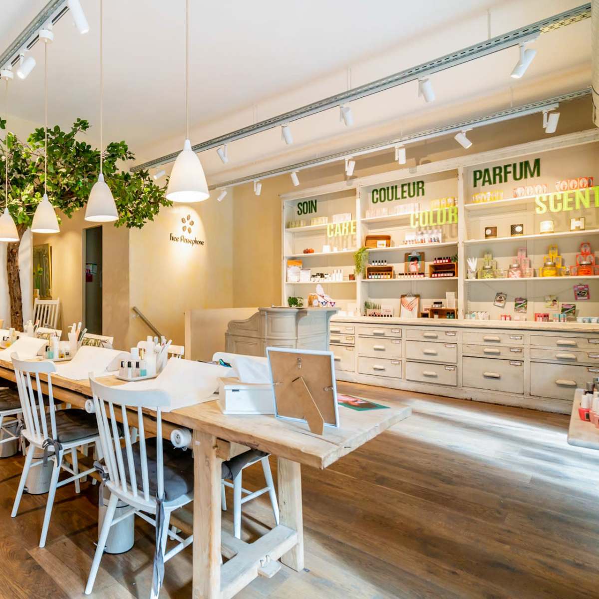 A spacious and well-lit interior features a wooden communal table surrounded by white chairs, with shelves displaying various products in the background.