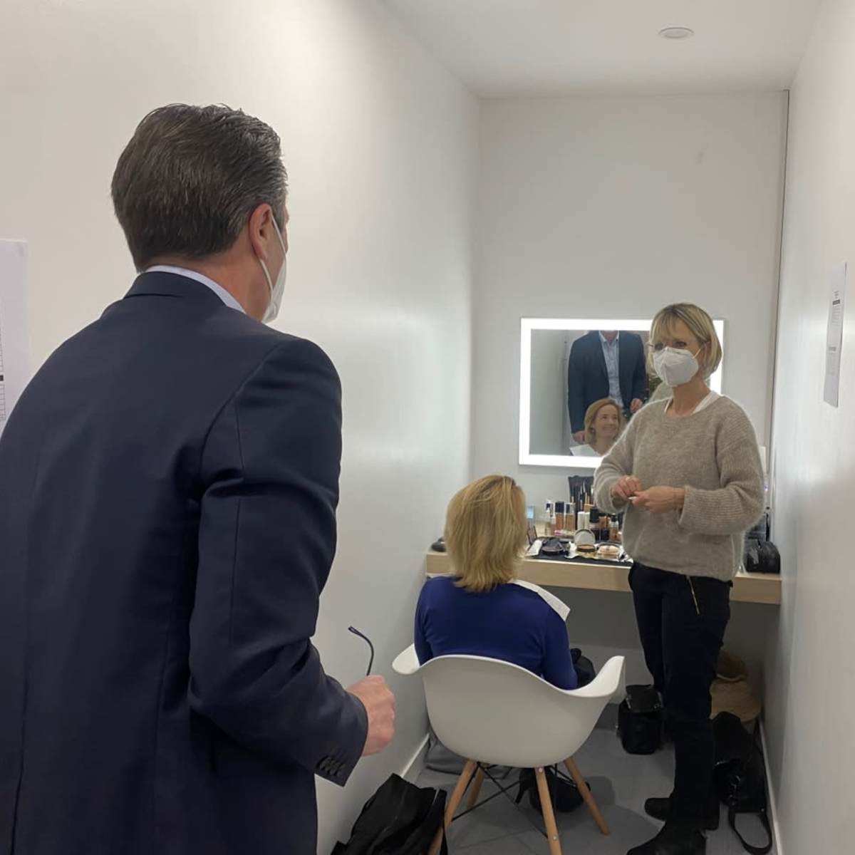 A man in a suit stands talking to a woman applying makeup to another woman seated in front of a mirror in a brightly lit room.