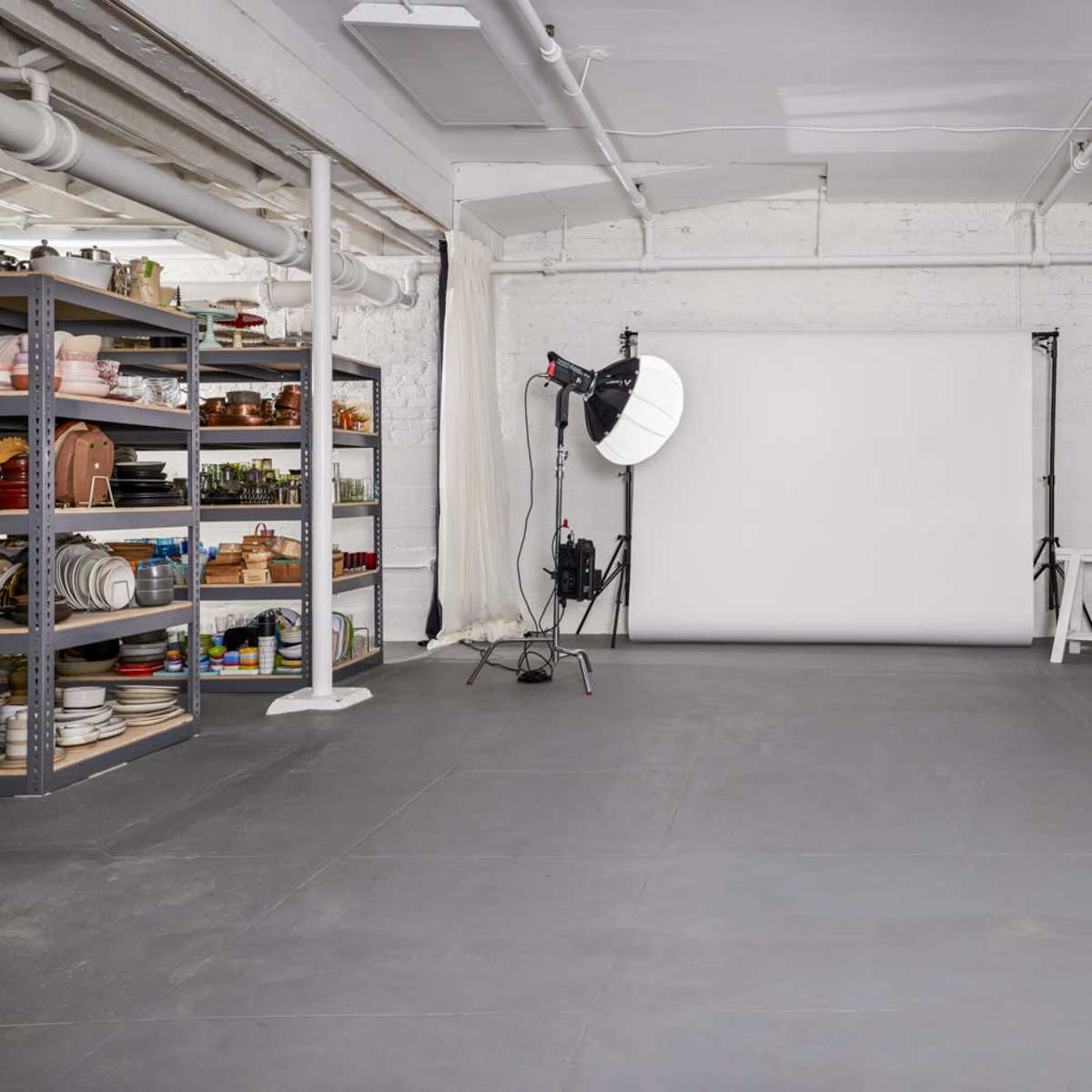 A spacious photography studio with a blank backdrop, lighting equipment, a shelving unit filled with various props, and a large mirror.