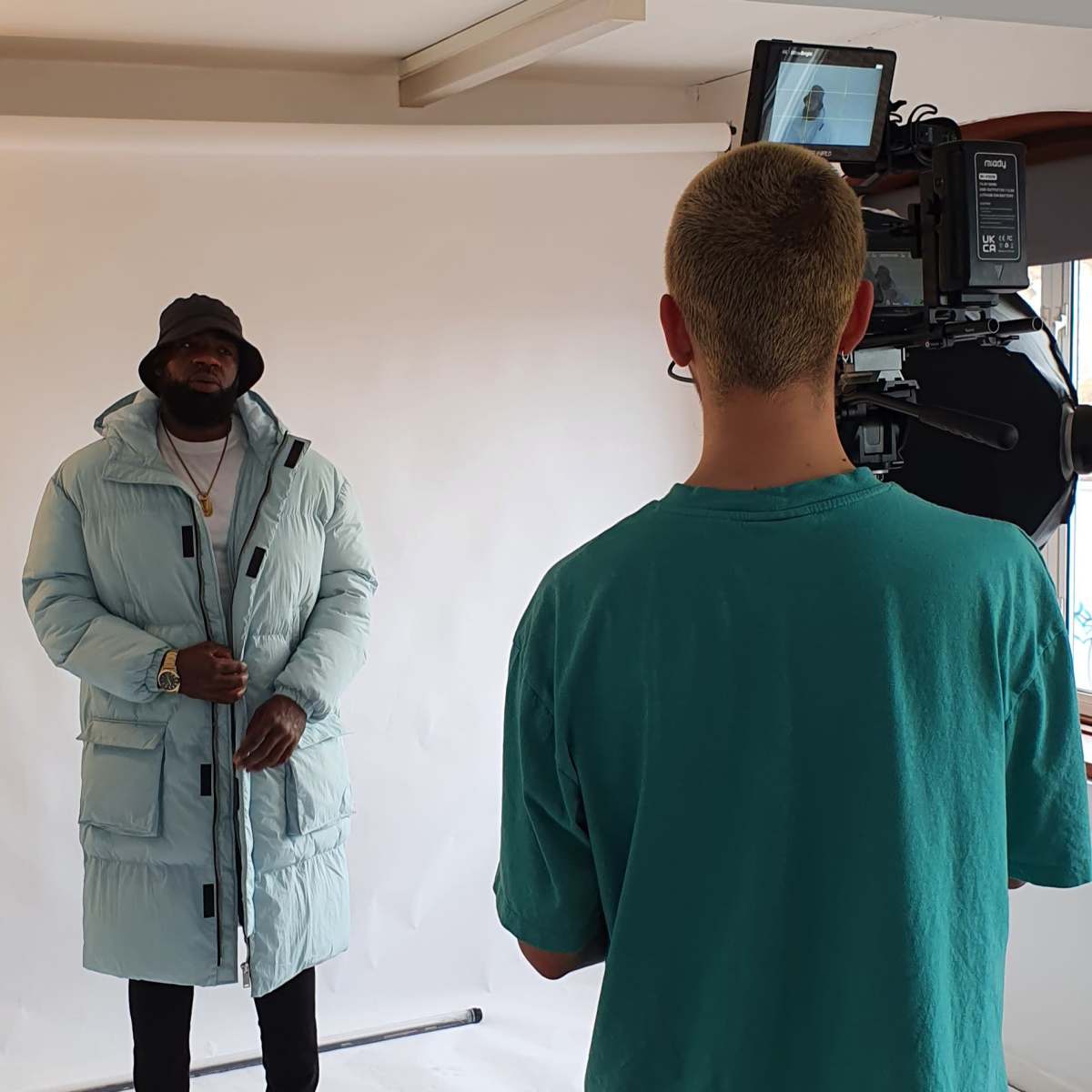 A man in a light blue puffer coat stands in front of a camera, while a crew member operates the equipment from behind.