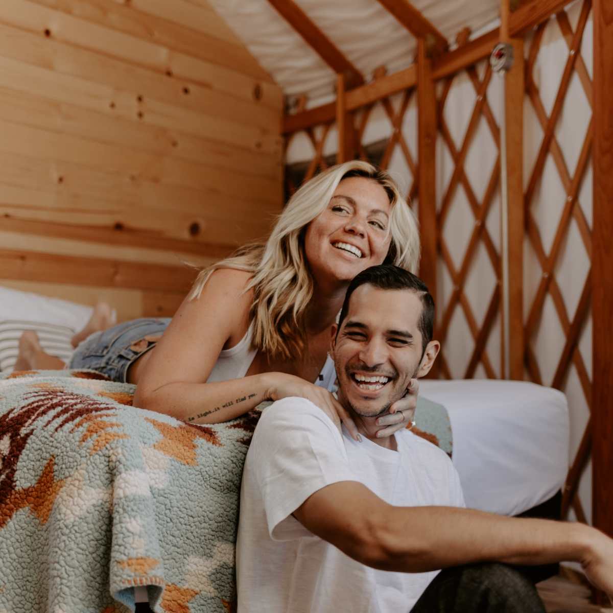 A woman sits on a bed smiling at a man who is seated on the floor, both in a cozy cabin interior.