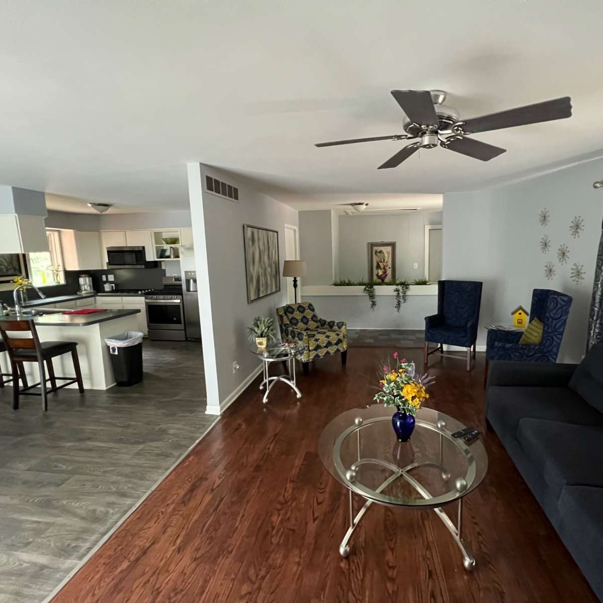 A modern living room area with a gray sofa, a glass coffee table, and two blue chairs, adjacent to a kitchen space with black appliances and a dining area.