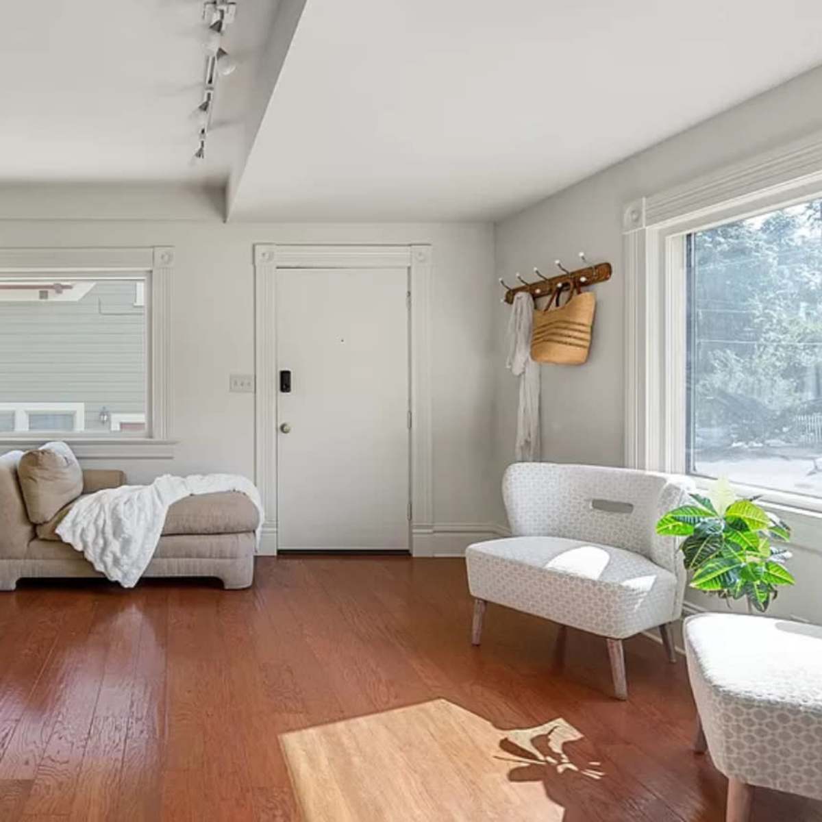 A bright room with a light-colored couch draped in a blanket, a glass display cabinet, and a small chair beside a window with a plant on the floor.