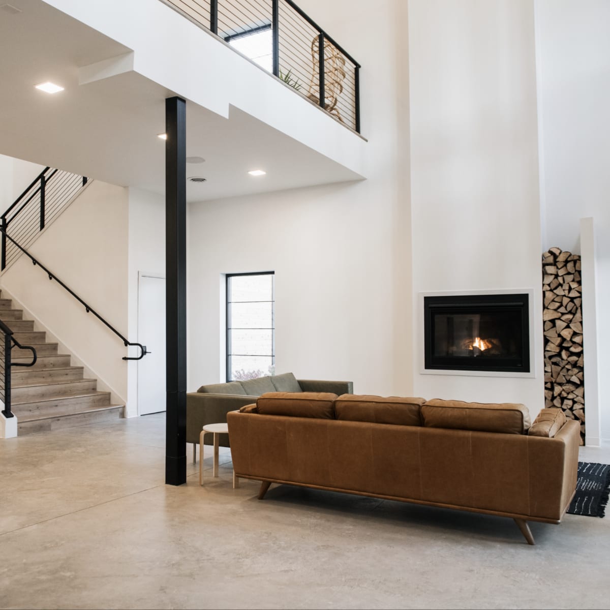 A modern interior with a staircase, a seating area featuring a brown sofa, and a fireplace with stacked wood nearby.
