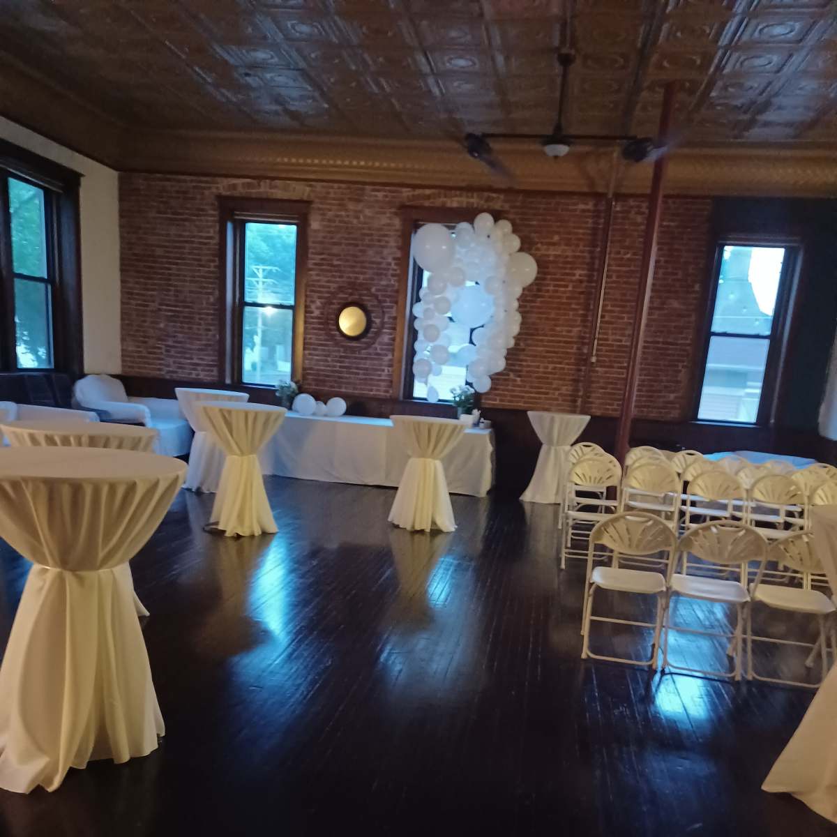 An interior venue set up for an event, featuring white tablecloths on high tables, chairs arranged in rows, and a decorative balloon installation against the brick walls.