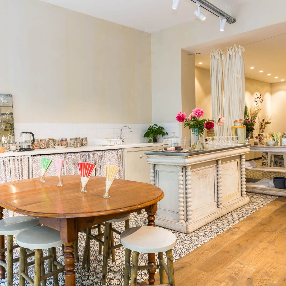 A bright, airy kitchen and dining area with a wooden table, pastel-colored chairs, and a counter lined with jars and flowers.