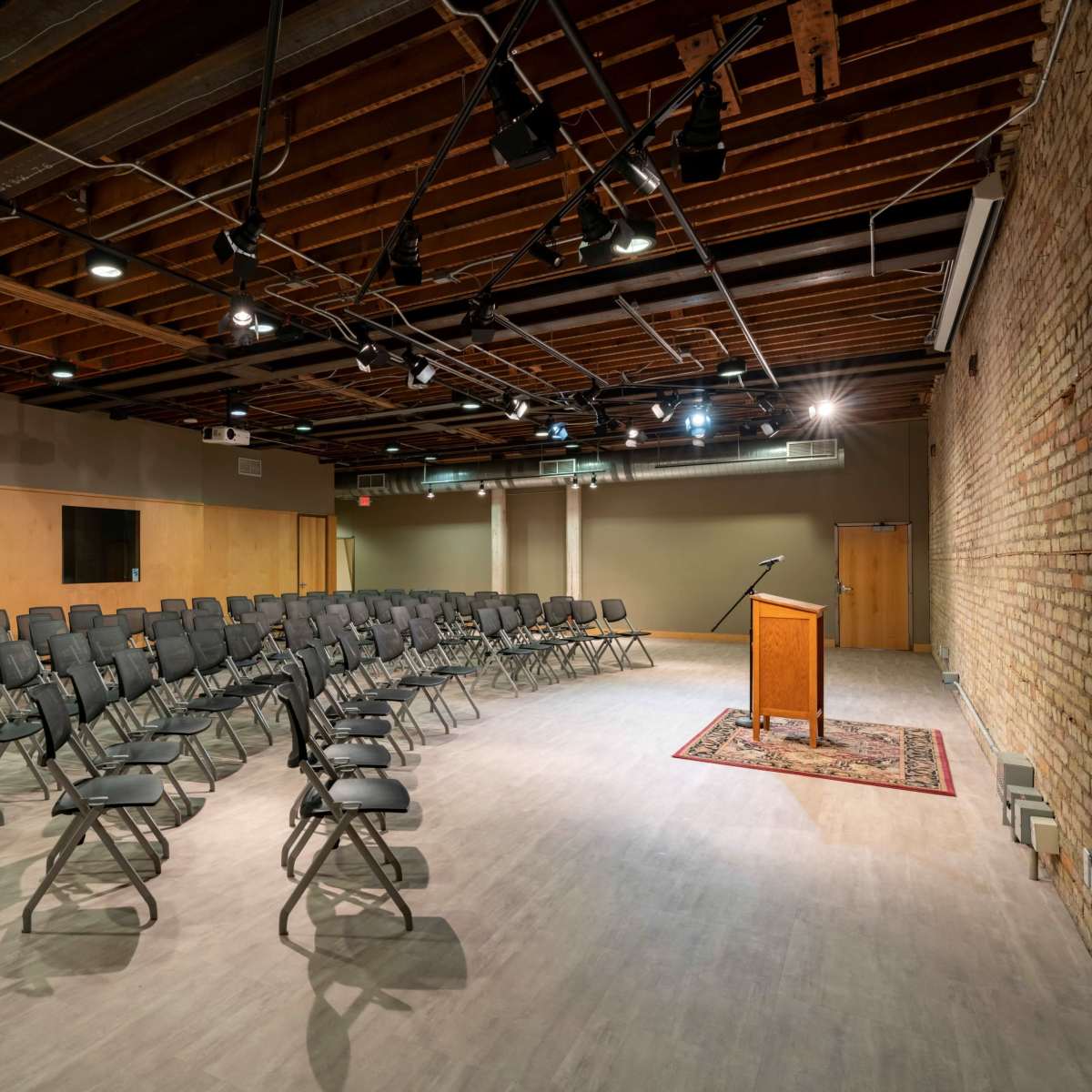 A spacious room with rows of black folding chairs arranged facing a wooden podium on a rug, with exposed brick walls and overhead lighting.