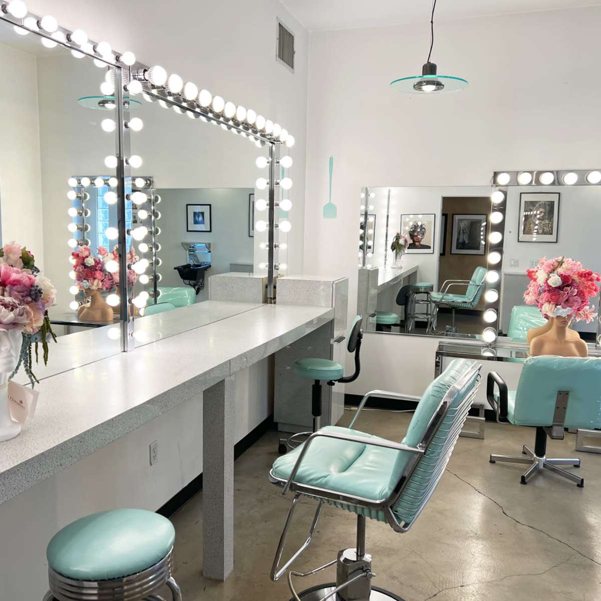 A brightly lit hair salon with a large mirror, a marble countertop, turquoise chairs, and floral arrangements placed on the counters.