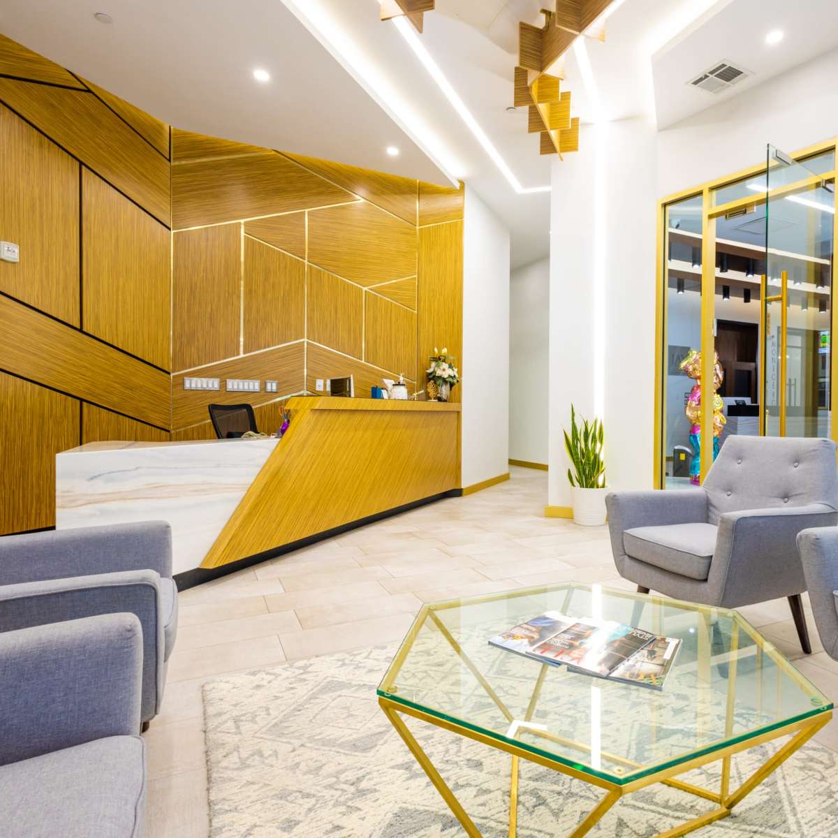 A modern reception area with a wooden reception desk, gray armchairs, and a glass table on a patterned rug.