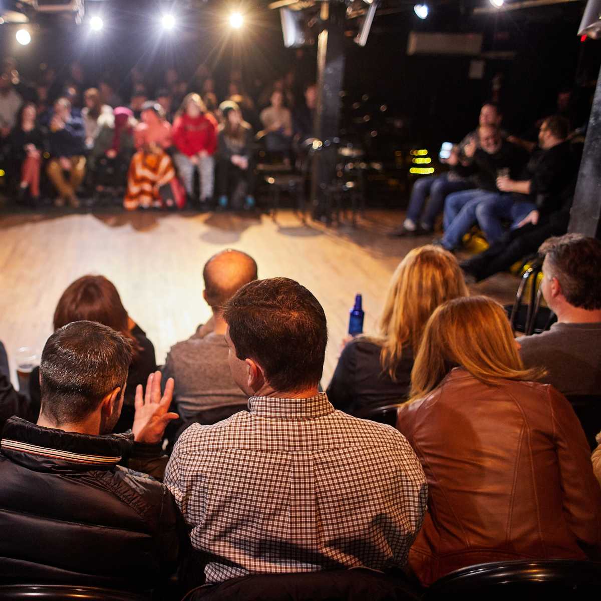 A group of people sits in a black box theater, facing a performance area illuminated by spotlights.