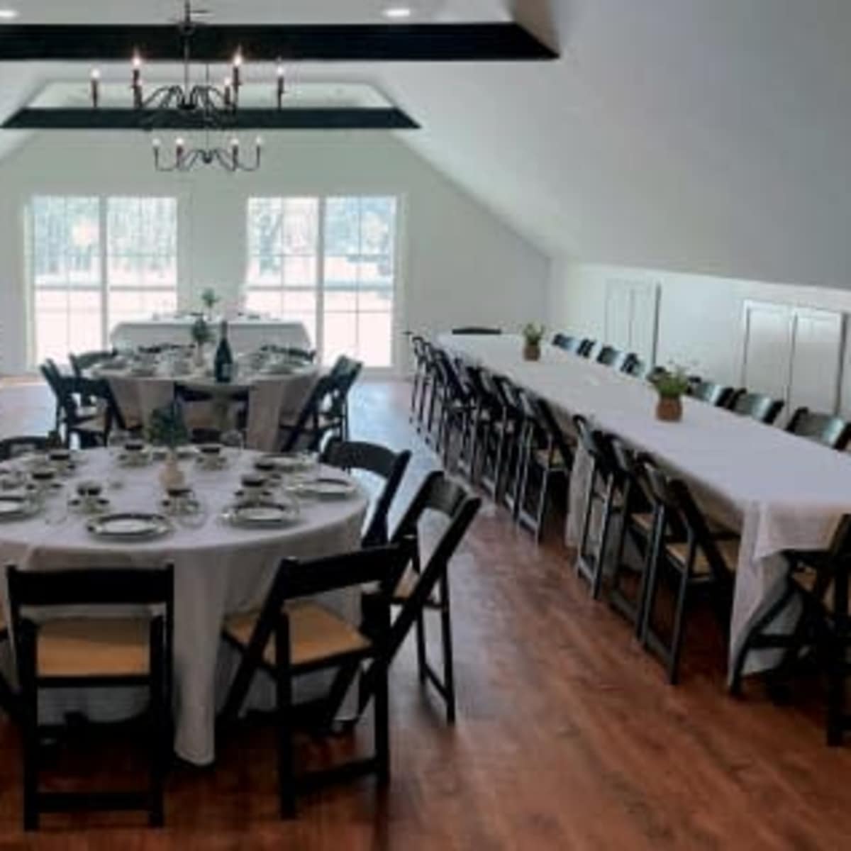 A spacious dining area features long tables set for a meal, with a chandelier hanging from the ceiling and large windows providing natural light.