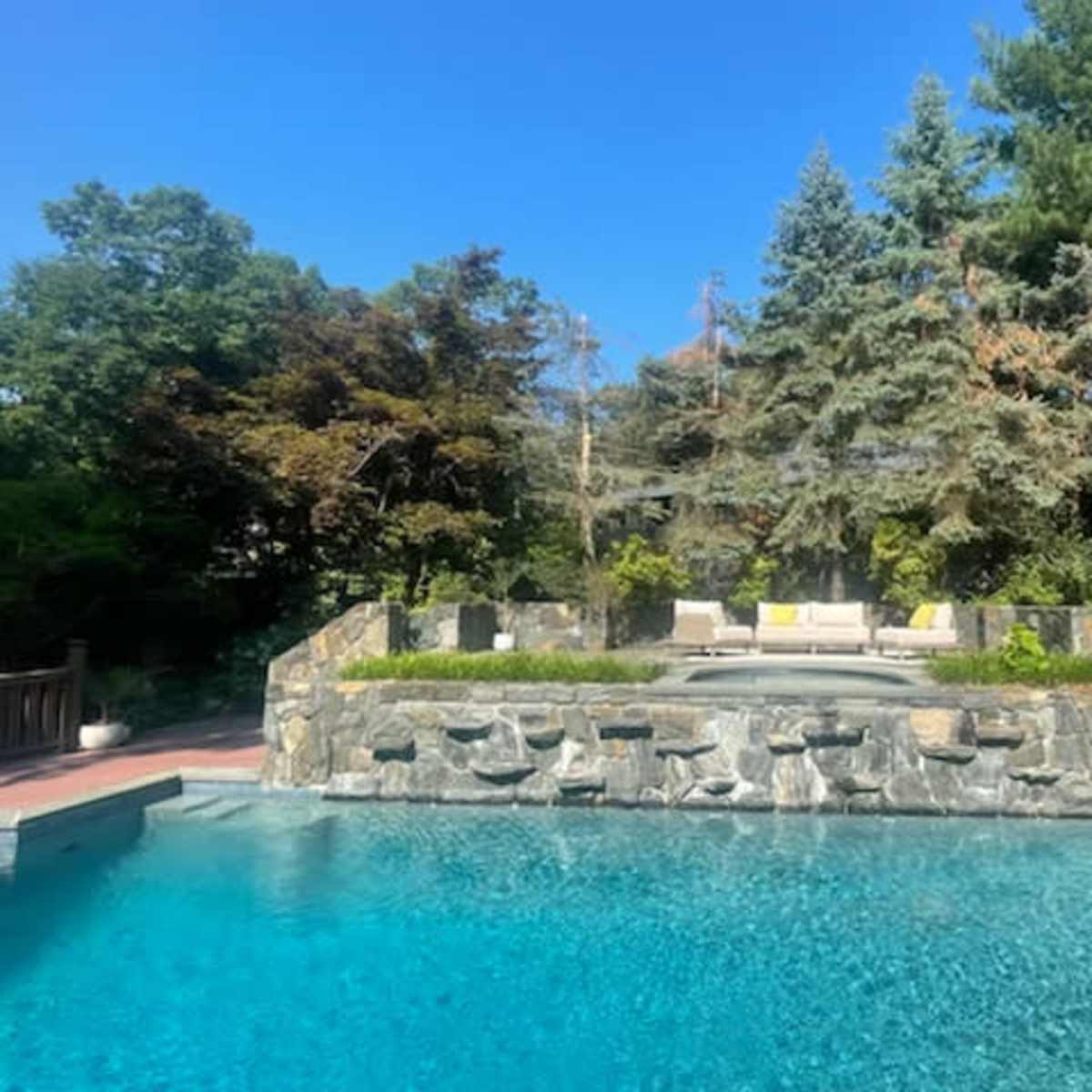A pool surrounded by a stone wall and landscaped gardens, with a seating area visible in the background.