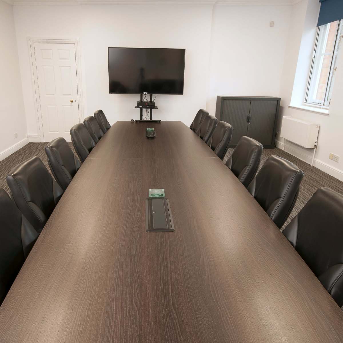 A large conference room features a long wooden table surrounded by black leather chairs, with a television mounted on the wall.