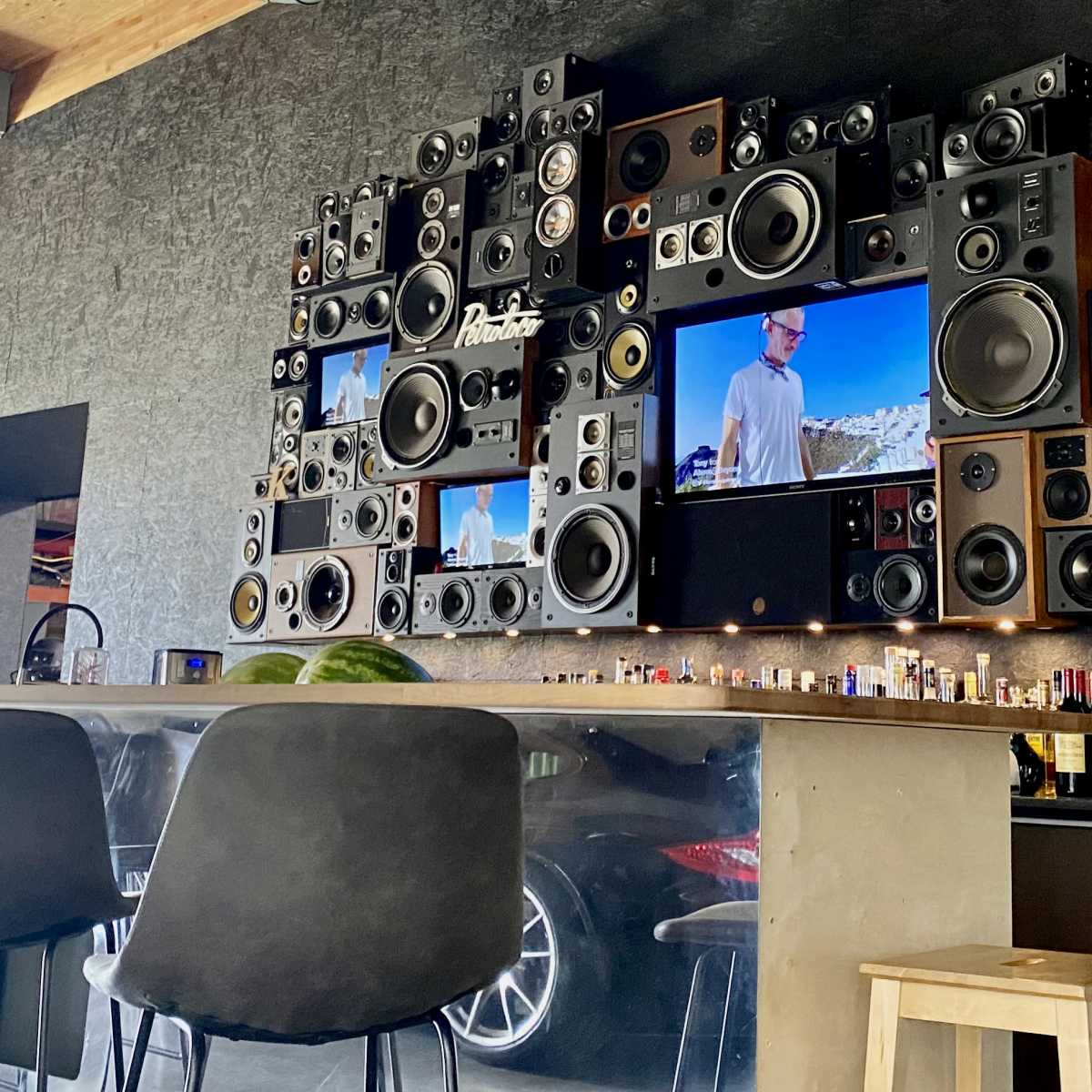 A wall-mounted arrangement of speakers and televisions in a modern bar area with a concrete counter and high stools.