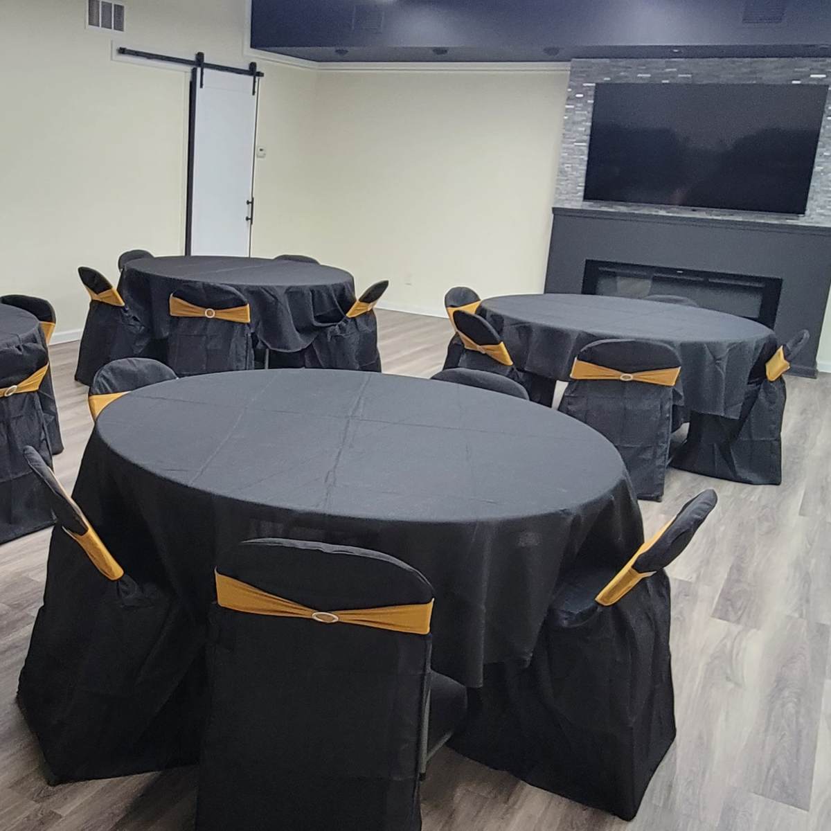 A room set up for an event, featuring several round tables covered with black tablecloths and decorated with gold chair sashes.