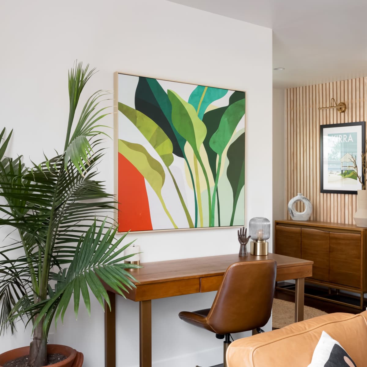 A modern living space features a desk with a brown chair, a large green and orange artwork on the wall, and a mirrored door leading to another room.