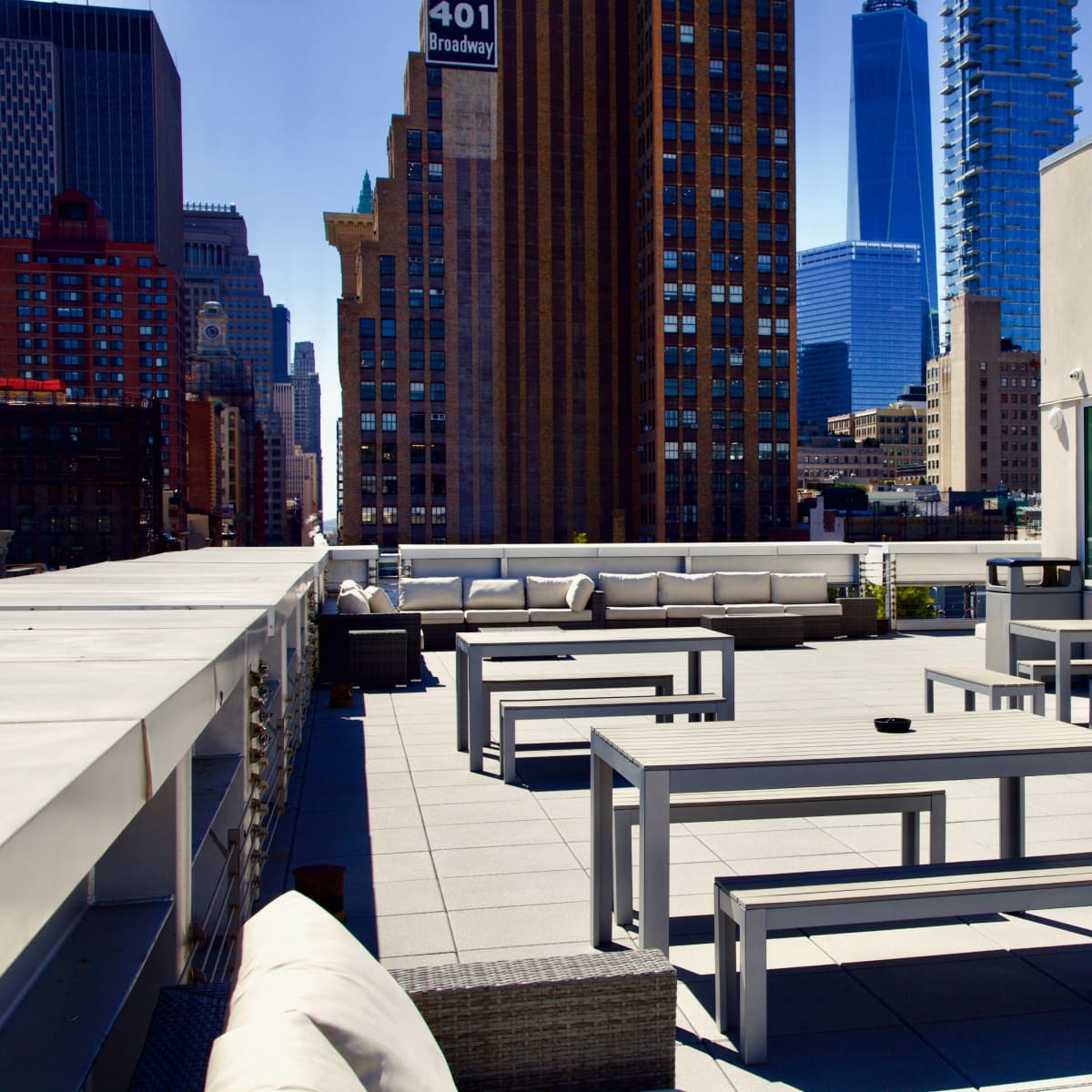 A rooftop terrace with several gray tables and benches, surrounded by tall buildings and a clear blue sky.