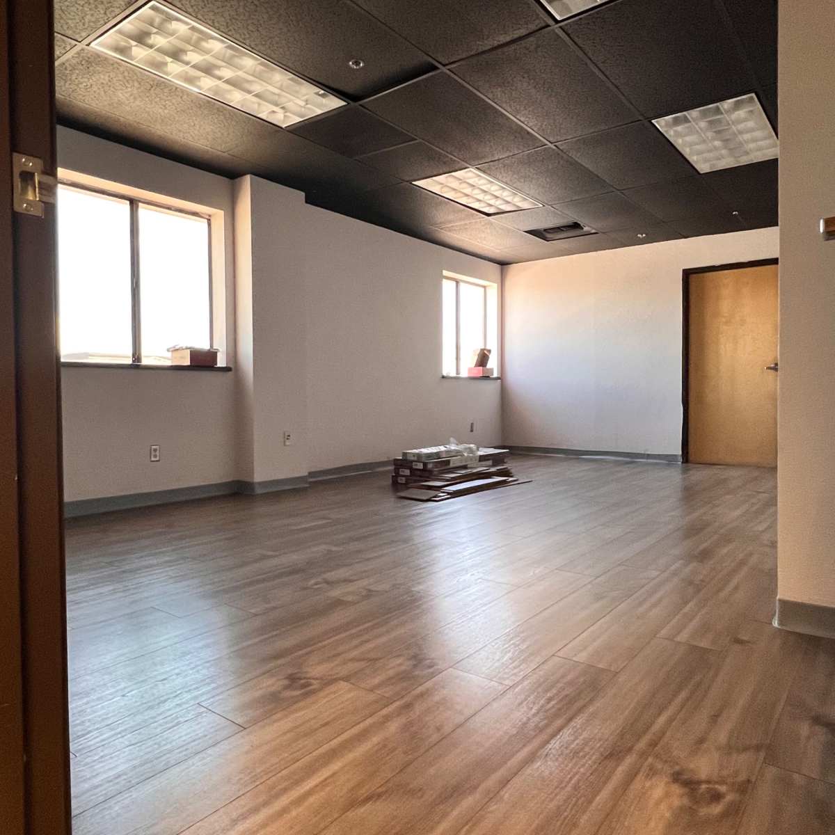 A vacant office space with wooden floors, two windows, and an open door leading into the room.