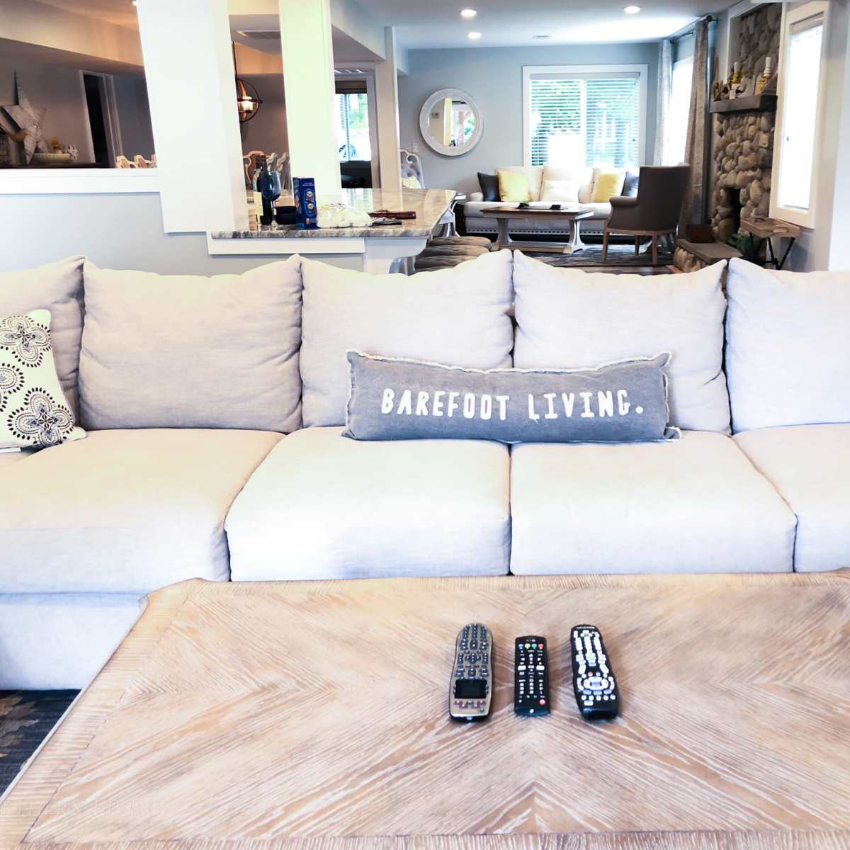 A light gray sectional sofa with decorative pillows is centered in a modern living room, accompanied by a wooden coffee table displaying remote controls.