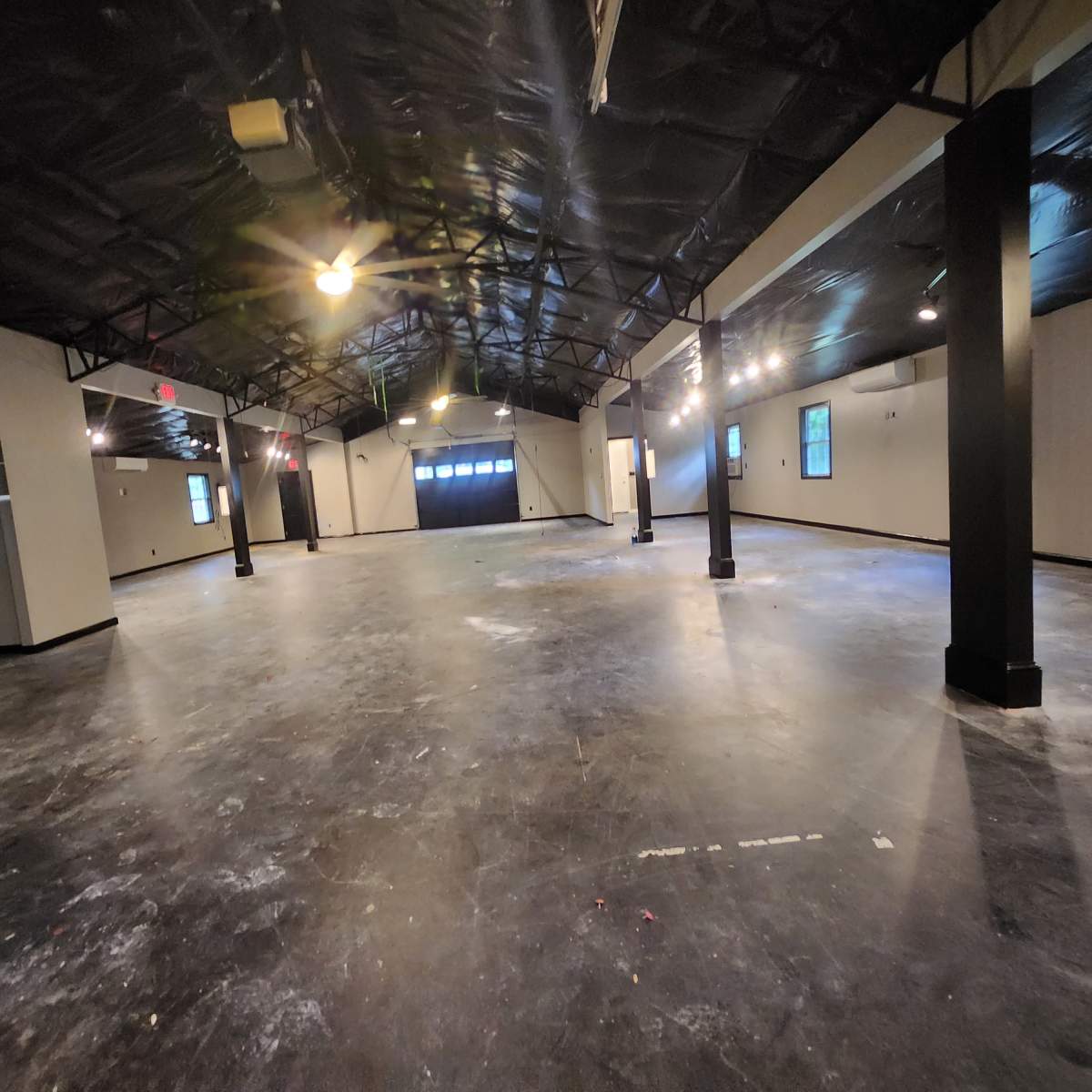 A spacious, empty room with a high ceiling, black pillars, and visible windows along the sides.